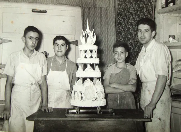 Fotografia antiga en blanc i negre: quatre pastissers amb davantal al costat d'un pastís de noces, a l'obrador de Pastisseria Miró